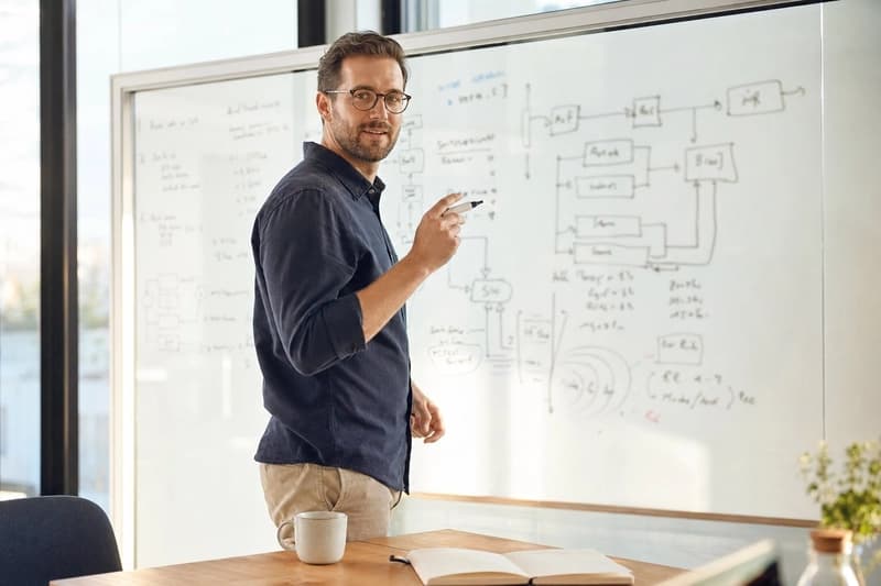 An engineer gesturing toward a system design diagram sketched on a glass whiteboard during an interview.